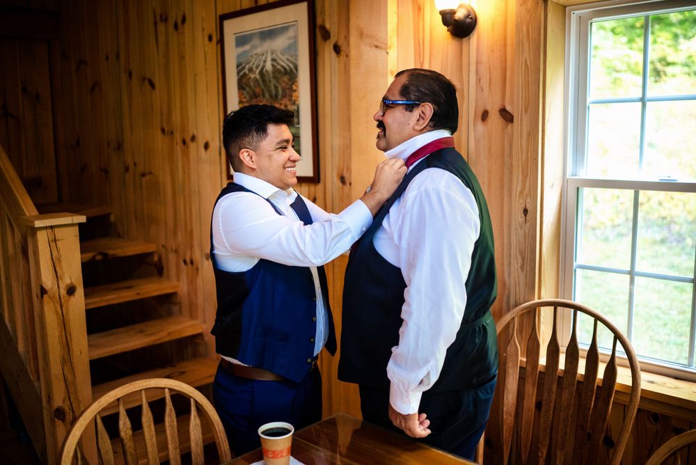 Groom adjusts groomsman tie during wedding day preparations in a rustic wooden cabin.