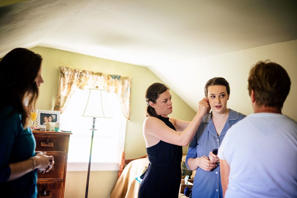 Bridesmaids help bride with earrings during wedding morning preparations in a cozy room.