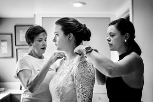 Two women help bride fasten her lace wedding dress during bridal preparations in black and white.