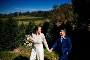 Smiling bride and groom hold hands while strolling through an evergreen tree farm on their wedding day.
