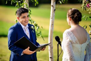 Groom smiles joyfully while reading wedding vows beneath a birch tree during outdoor ceremony.