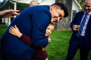 Groom in a blue suit joyfully hugging a young boy at a wedding celebration.