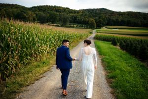 Bride and groom walking hand in hand down a gravel path beside a cornfield.