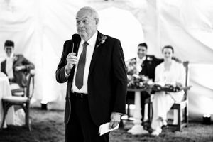 Older man delivers a heartfelt wedding speech into a microphone in black and white photo.