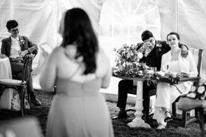 Groom wipes tears during wedding speeches while bride smiles beside him, black and white.