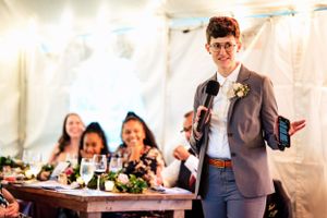 Person in gray blazer delivers a wedding toast holding a microphone and phone.