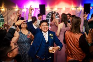 Groom in blue suit celebrates on the dance floor with guests under colorful lights.