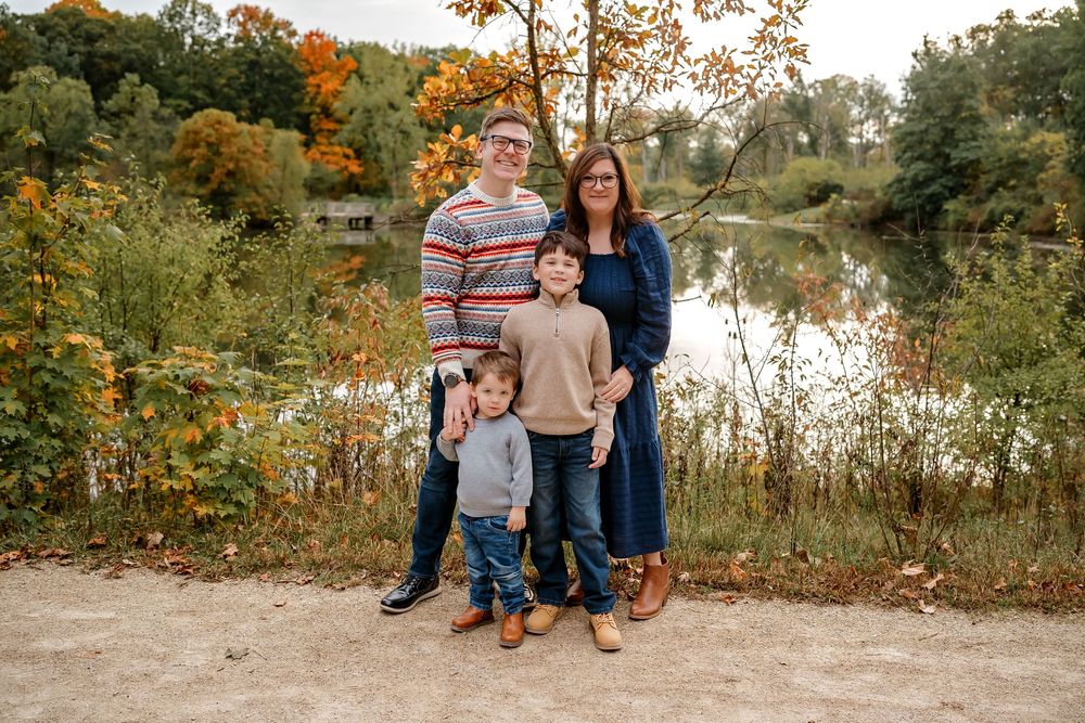 Family in coordinating fall outfits poses together by a lake during autumn with colorful foliage in the background.