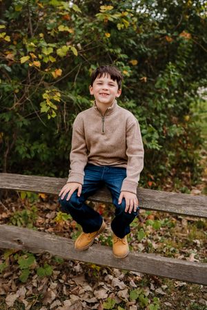 A young child sits on a rustic wooden fence surrounded by autumn foliage wearing a brown sweater and blue pants.
