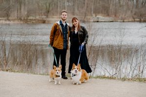Couple walking two Corgi dogs on leashes near a lake in a park during early spring.