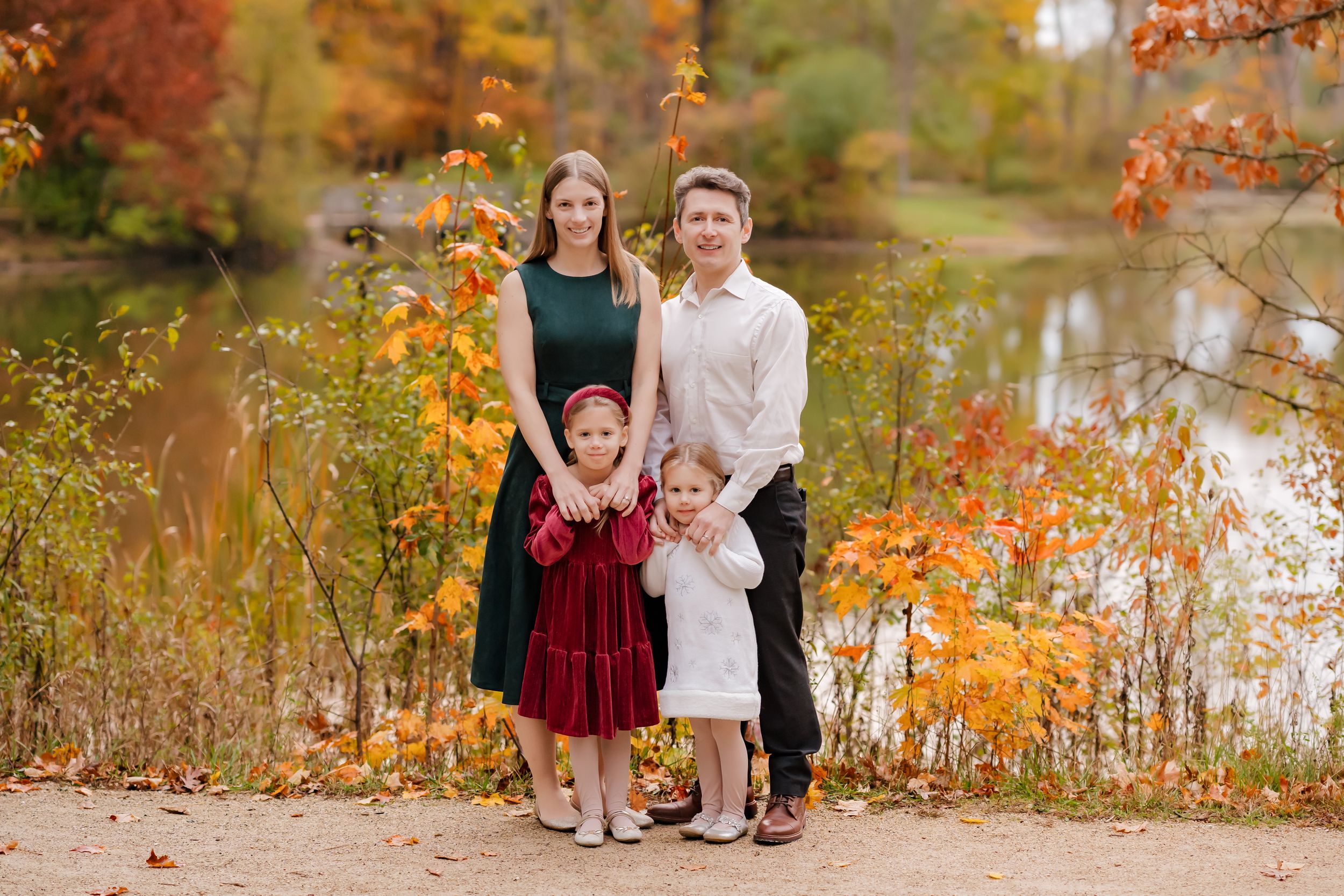 Family group portrait in autumn setting with colorful fall foliage and lake in background.