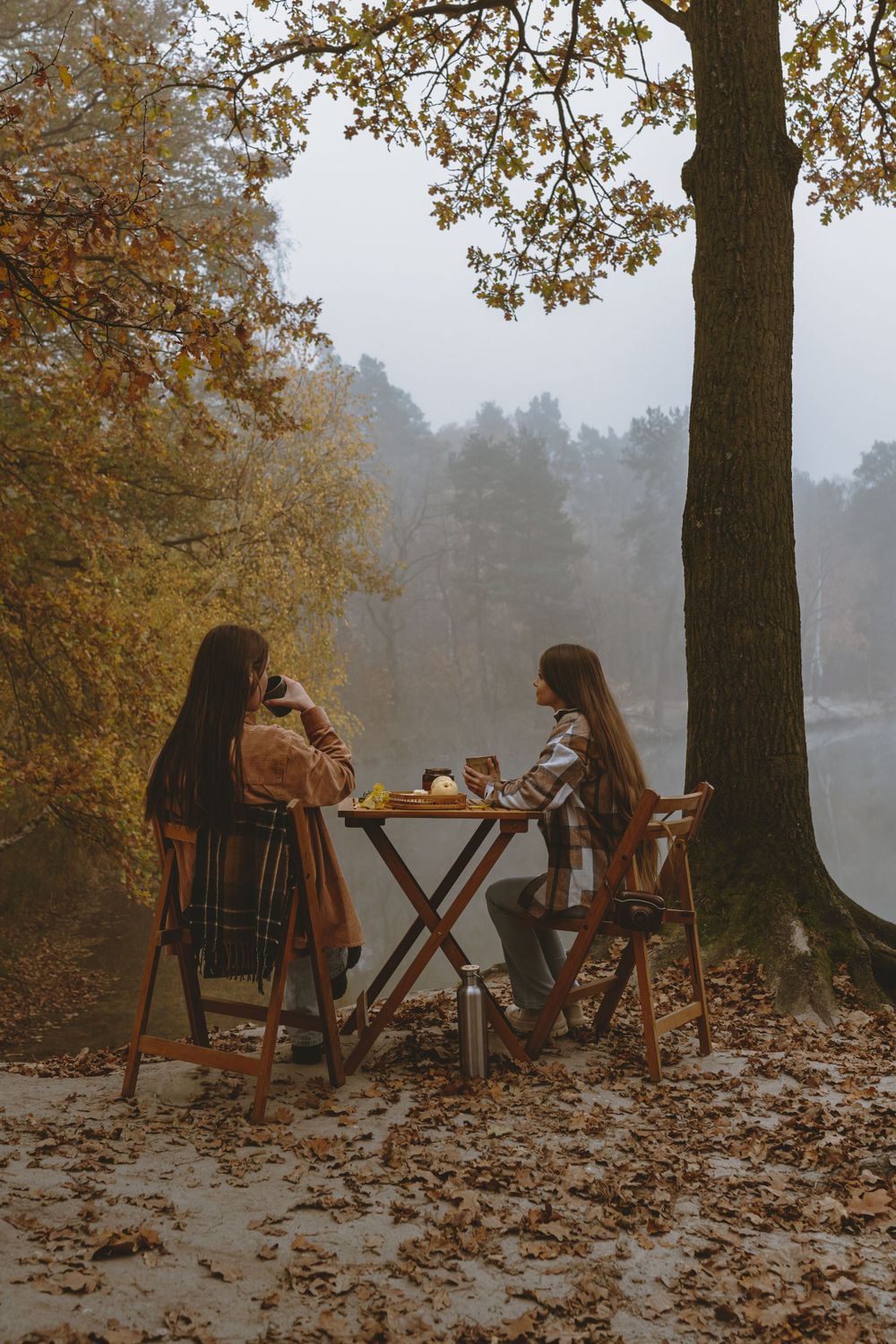 Two people enjoying tea at wooden table by misty autumn lake.