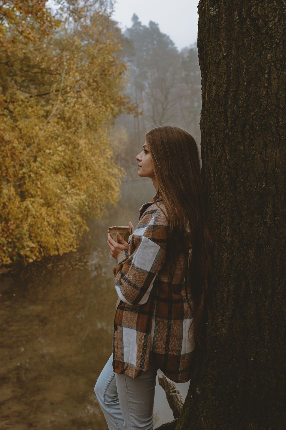 Person in plaid shirt leaning against tree trunk in autumn forest.