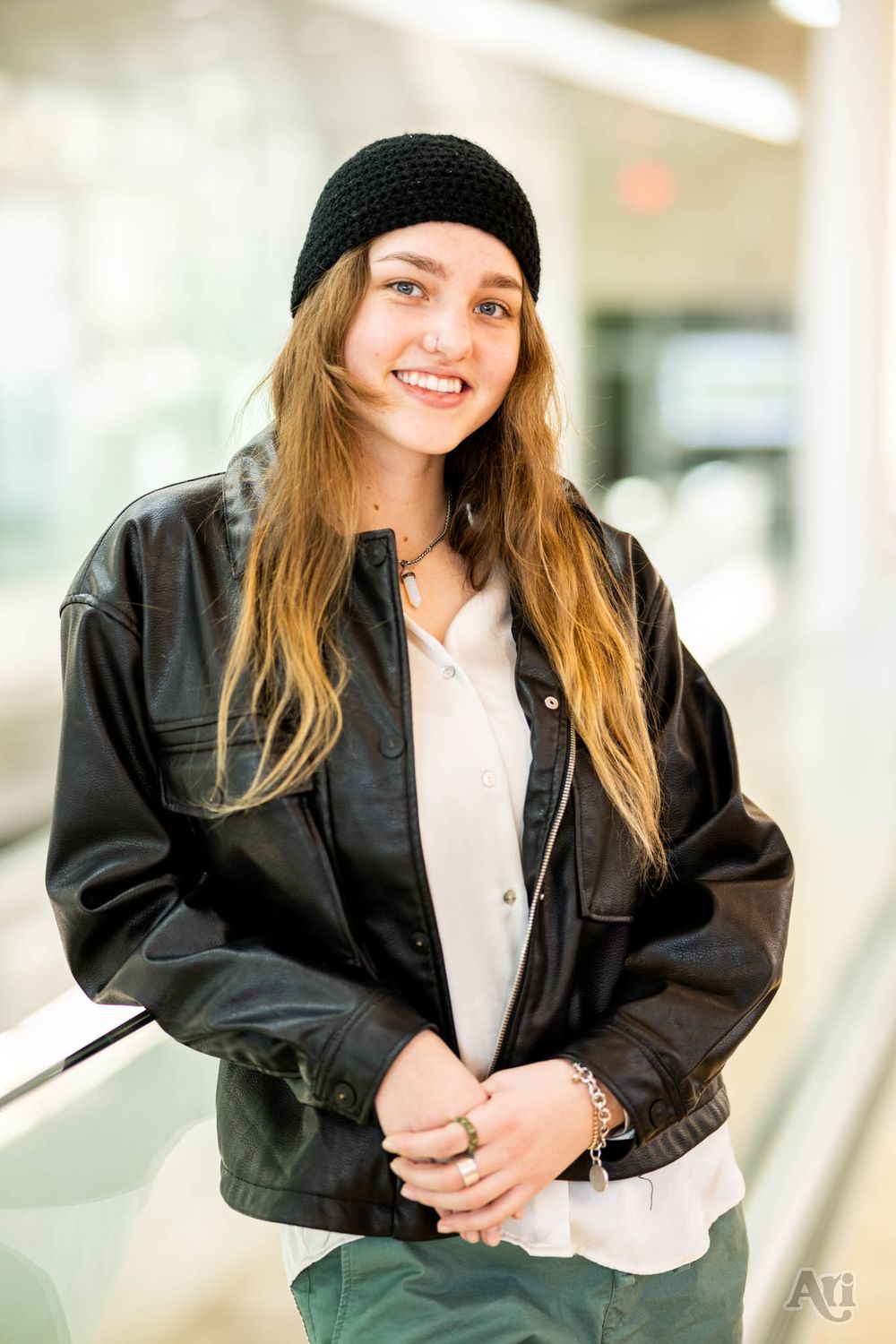 A casual portrait of someone in a black knit cap and leather jacket standing in a bright indoor setting.
