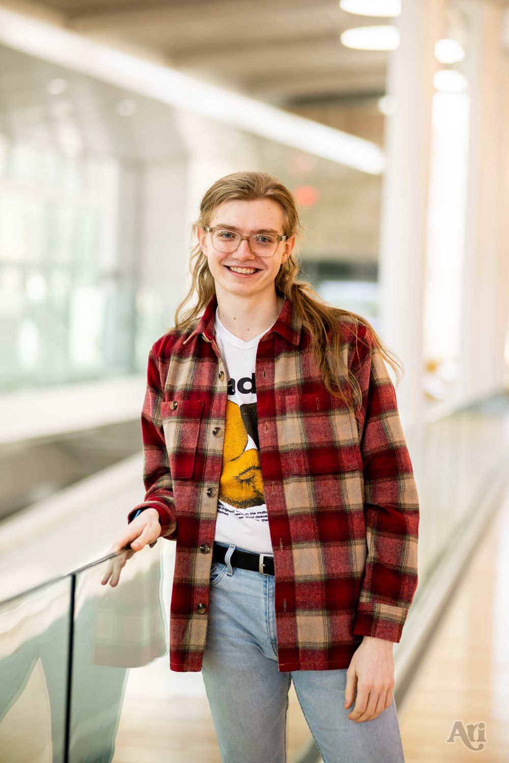 A portrait of someone wearing glasses and a red plaid flannel shirt in a bright indoor hallway.