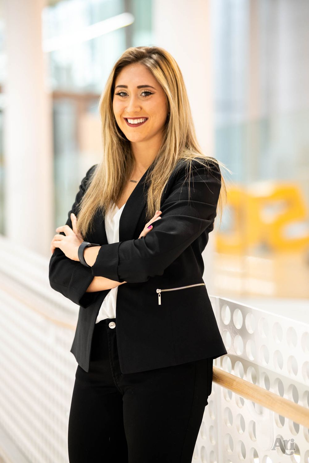 A professional portrait of someone in a black business suit standing with crossed arms in a bright hallway.