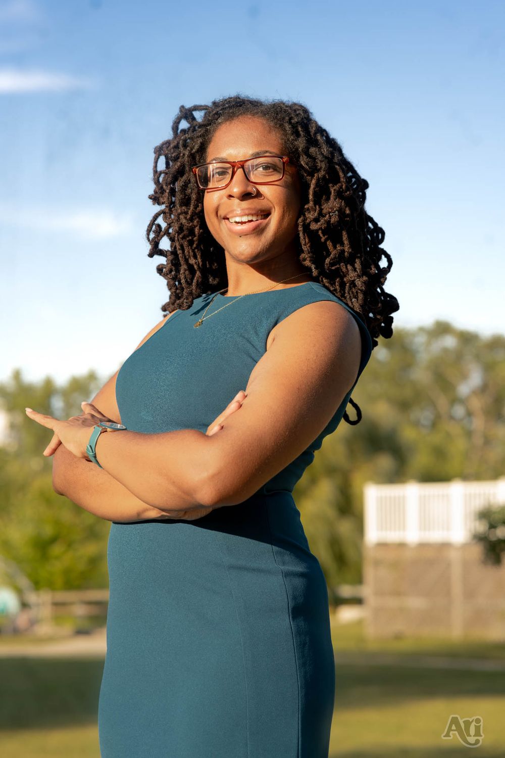 A portrait of someone in a teal dress standing outdoors with crossed arms against a nature background.