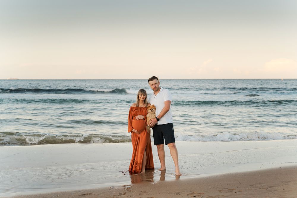 Couple stands together on a sandy beach at sunset as ocean waves roll in behind them during a pregnancy photoshoot in Sydney Maroubra Beach