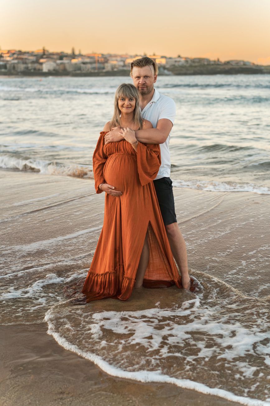 A couple on a sandy beach at sunset as gentle waves wash around them in a romantic seaside moment during a maternity photoshoot in Maroubra
