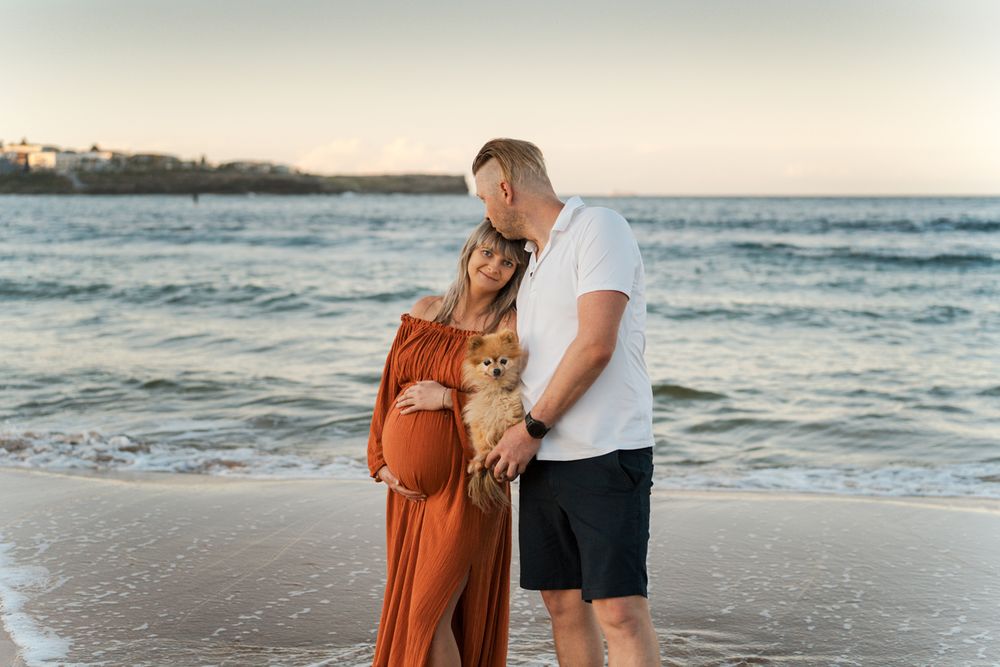 Couple and their dog share a moment on the beach at sunset with waves rolling in the background during their maternity photoshoot in Sydney