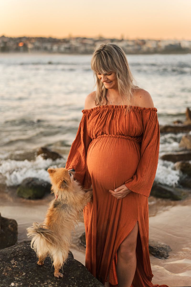 A mom to be in a rust-colored dress stands on a beach at sunset with a small fluffy dog beside her during a maternity photoshoot in Sydney
