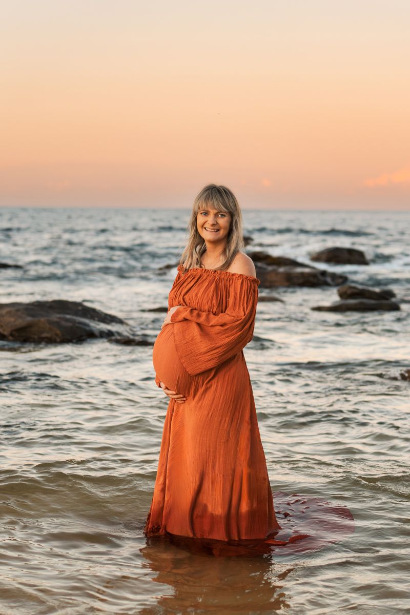 Expecting mother in flowing rust colored dress stands in ocean water at sunset with rocky coastline in background maternity photoshoot Sydne