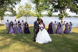 Wedding party poses together on grassy lawn by waterfront in purple and white formal attire.