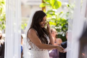 A bride in a white lace wedding dress laughs while holding a piece of paper during an outdoor ceremony.