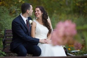 A couple in wedding attire sits close together on a park bench surrounded by autumn foliage and pink flowers.