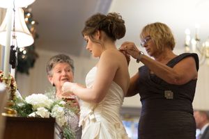 A bride gets help with her dress while standing near white floral arrangements and elegant chandeliers.