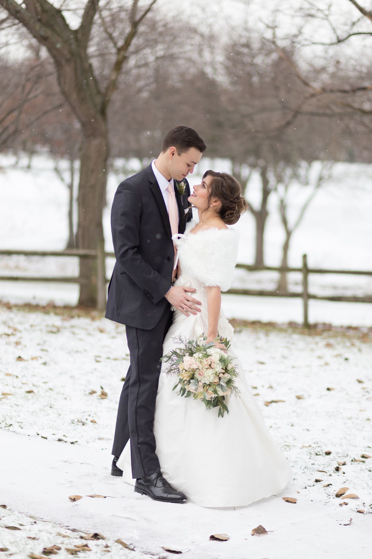 A winter wedding portrait with a couple standing in the snow with bare trees and a white fence in the background.