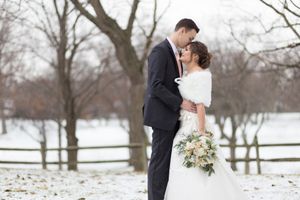 A couple shares a romantic winter moment outdoors with snow-covered trees and white fencing in the background.
