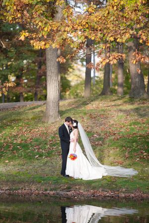 A romantic wedding couple stands by a pond surrounded by autumn trees with golden leaves and their reflection in the water.