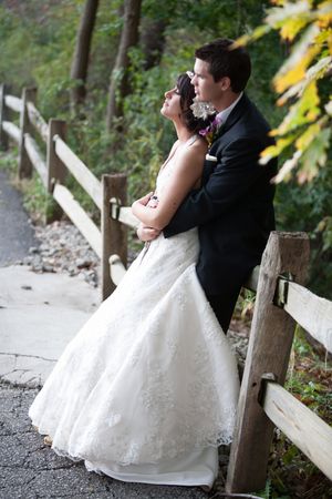 A couple in wedding attire embraces against a rustic wooden fence surrounded by fall foliage.
