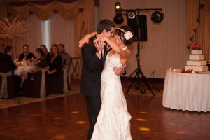 A couple shares their first dance at their wedding reception with a tiered cake visible in the background.