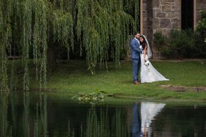 Romantic wedding couple poses by a serene pond with weeping willow trees in an elegant garden setting.