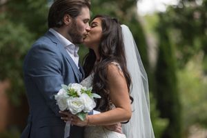 Newlywed couple shares romantic kiss in a lush garden setting while holding white floral bouquet.