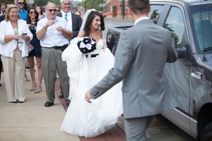 Wedding party and guests gather around a parked car during an outdoor celebration.