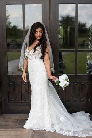 Elegant bride in a lace wedding gown with flowing cathedral veil poses against dark doors in an outdoor setting.