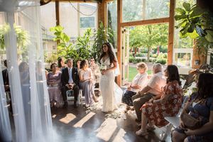 Sun-filled indoor wedding venue with guests seated along windows and natural greenery.