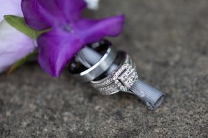 Close-up detail shot of wedding rings resting against purple flowers on a textured surface.