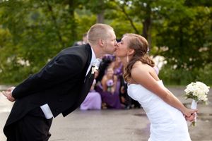 A romantic kiss between newlyweds in formal wedding attire surrounded by lush green trees in an outdoor setting.