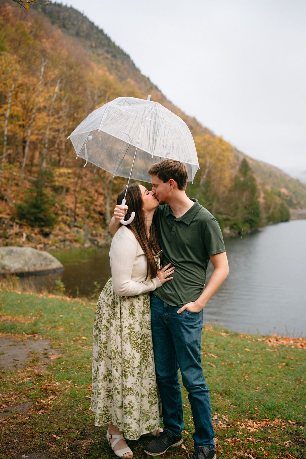 A couple shares a romantic moment under a clear umbrella beside a lake with autumn foliage in the background.