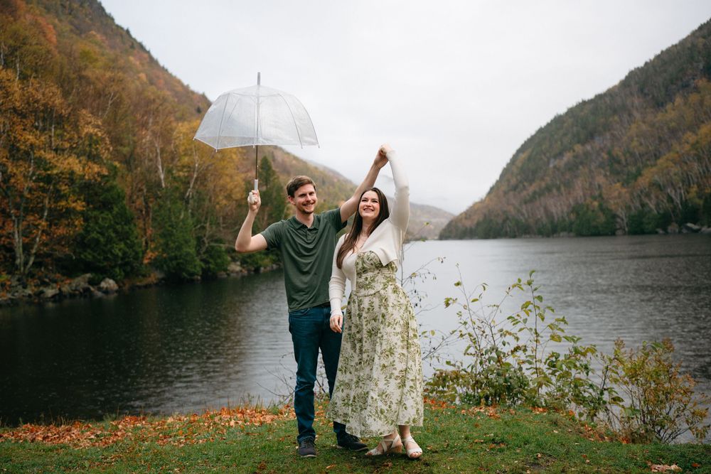 Two people share a white umbrella while celebrating by a scenic mountain lake on an autumn day.