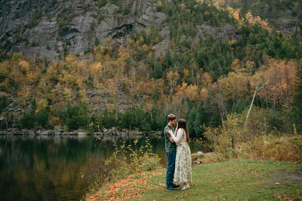A couple in formal attire stands by a serene lake surrounded by autumn foliage in a romantic outdoor setting.