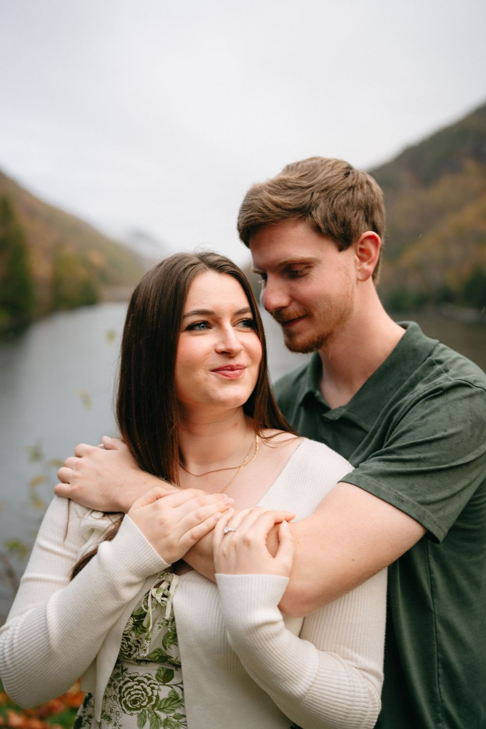 Romantic couple embraces by a scenic mountain lake during autumn with fall foliage in the background.