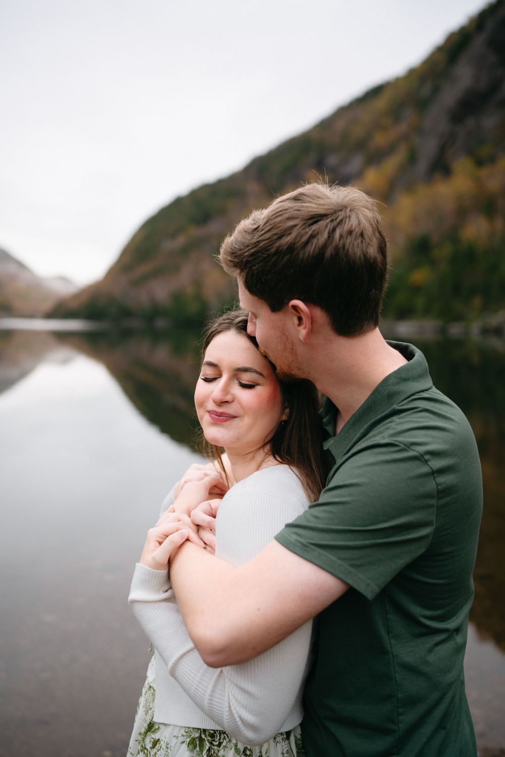 Couple in white and green share romantic moments by a tranquil mountain lake during fall season.