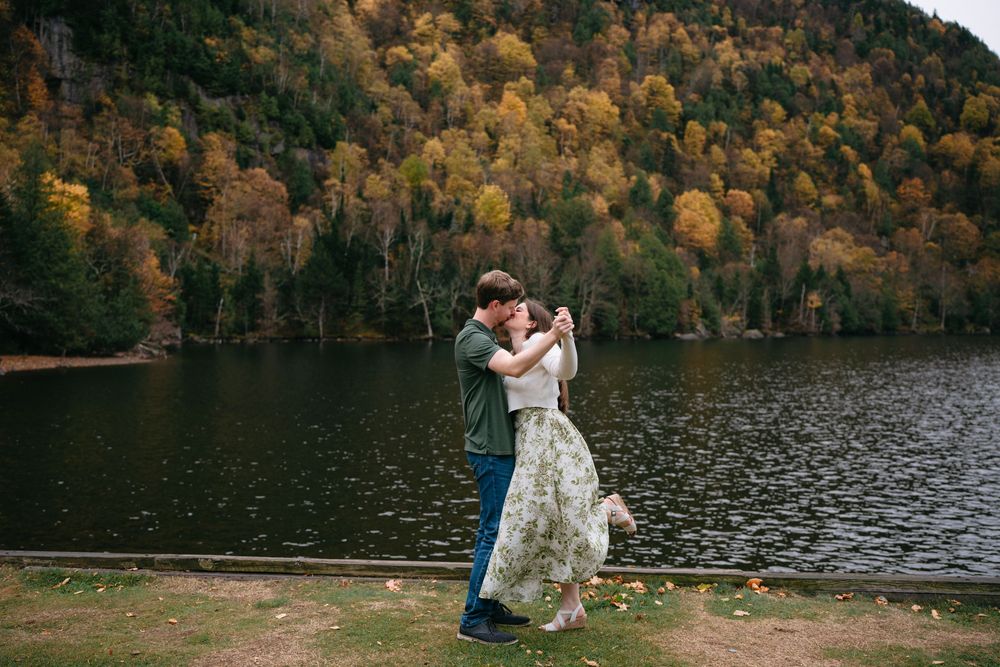 Couple embraces by a serene lake surrounded by autumn trees with golden foliage reflecting in the water.