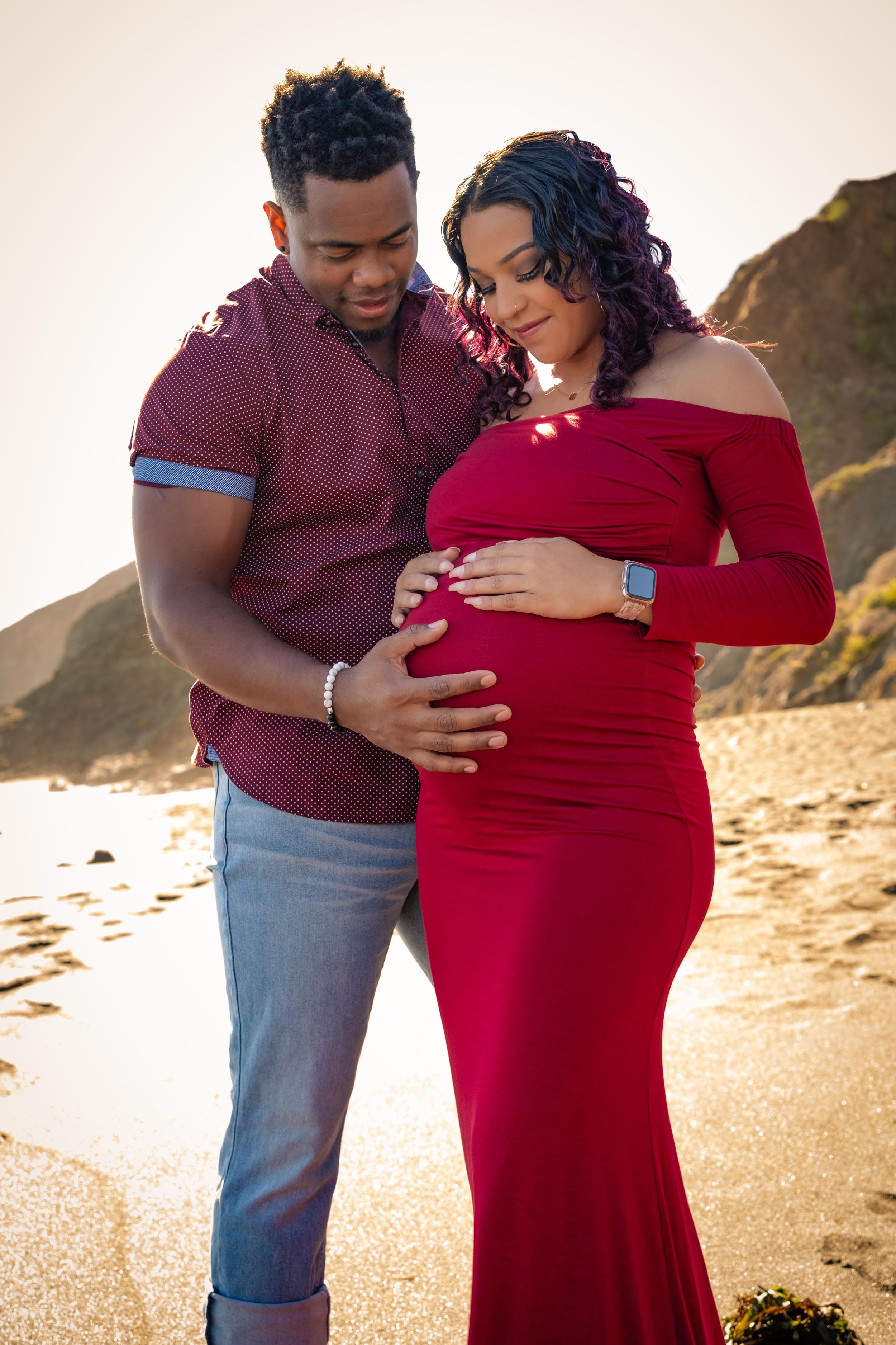 A couple embraces on a beach at sunset while wearing burgundy and red outfits during a maternity photoshoot.