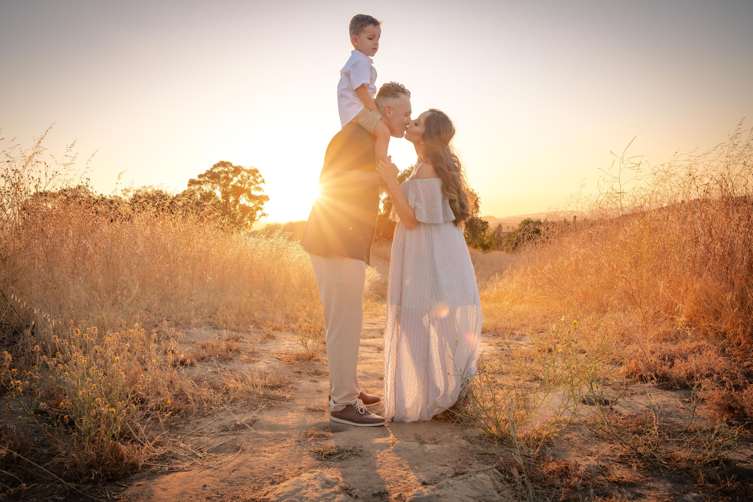 A couple embraces in a golden field during sunset as sunlight creates a warm, romantic glow through tall grass.